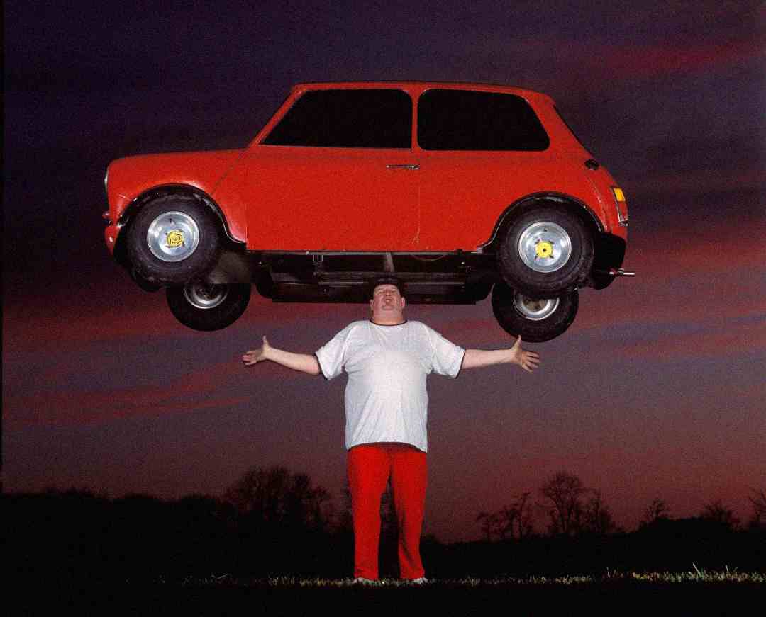 A person in a white shirt and red pants stands outdoors at sunset, holding a small red car above their head with both hands, as if effortlessly lifting it.