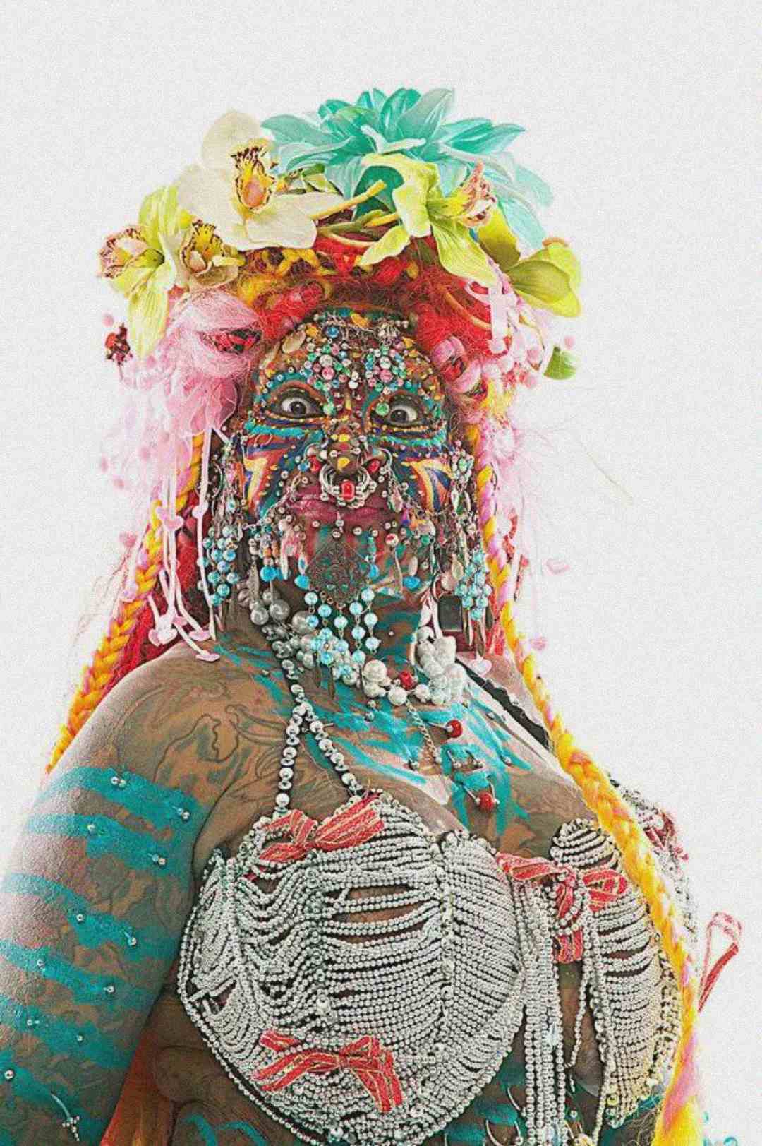 A person adorned with colorful face and body paint, beads, facial piercings, and wearing a vibrant headdress made of flowers and ribbons, along with a top decorated with pearls and silver chains.