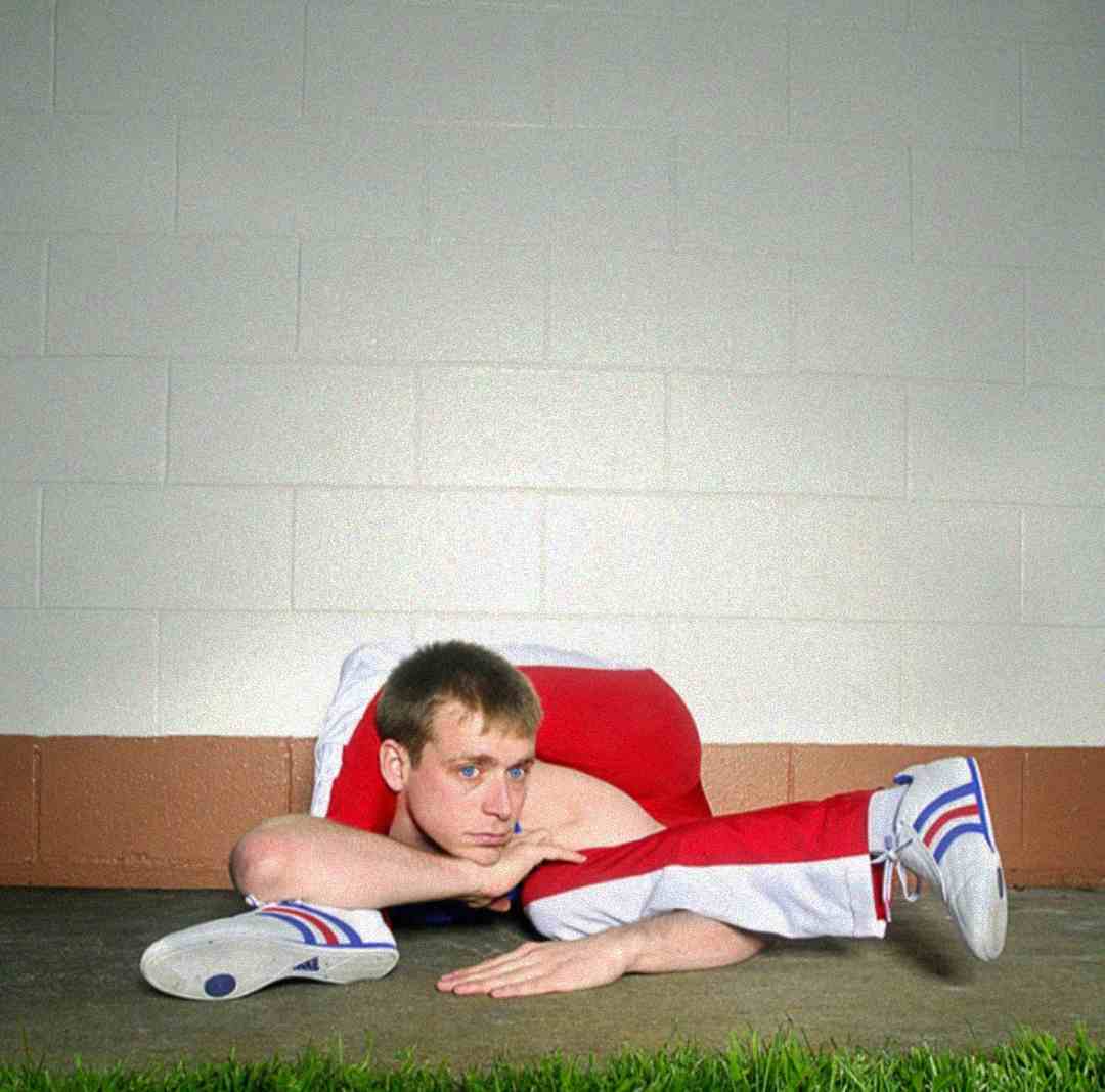 A man in red and white athletic clothing performs an extreme flexibility pose, resting his chin on his hand while his legs are bent over his shoulders, against a white brick wall with green grass in the foreground.