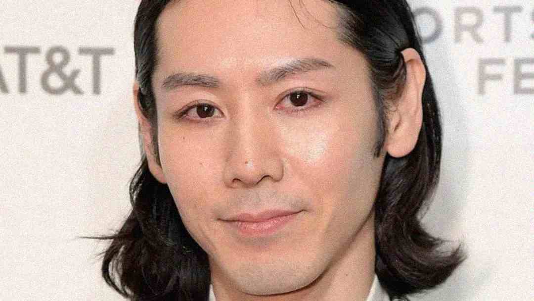 A person with medium-length dark hair and clear skin smiles softly at the camera, standing in front of a white background with partially visible text.