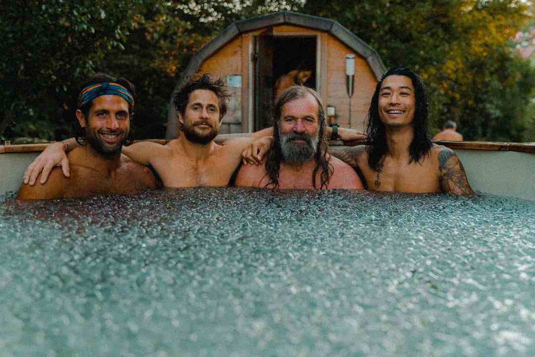 Four men with arms around each other smile while sitting in an outdoor hot tub, with a wooden sauna and greenery visible in the background.