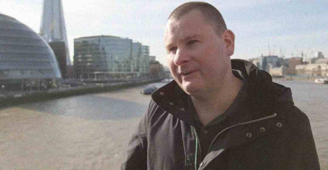 A man in a dark jacket stands by a river with modern glass buildings and the Shard skyscraper visible in the background on a clear day.
