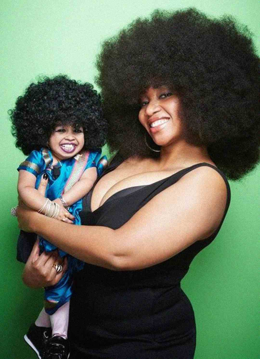 A smiling woman in a black dress holds a young child in a blue outfit. Both have large, curly afro hairstyles and pose in front of a green background.