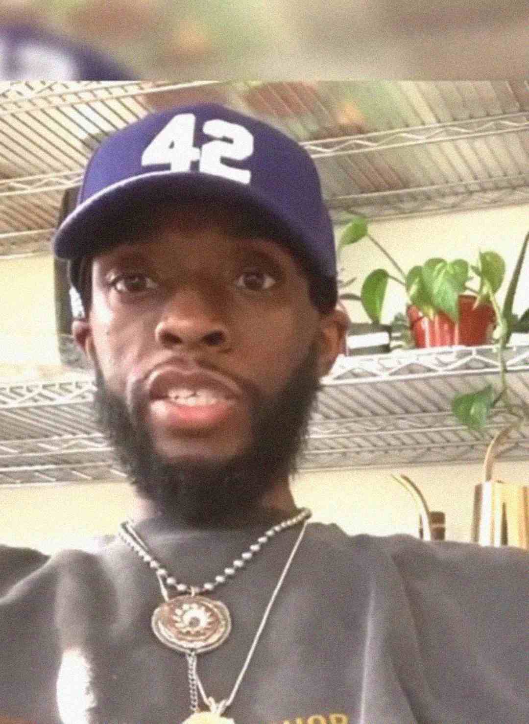 A man with a beard wearing a blue baseball cap with the number 42, a dark shirt, and necklaces sits indoors in front of metal shelves with plants and various items.