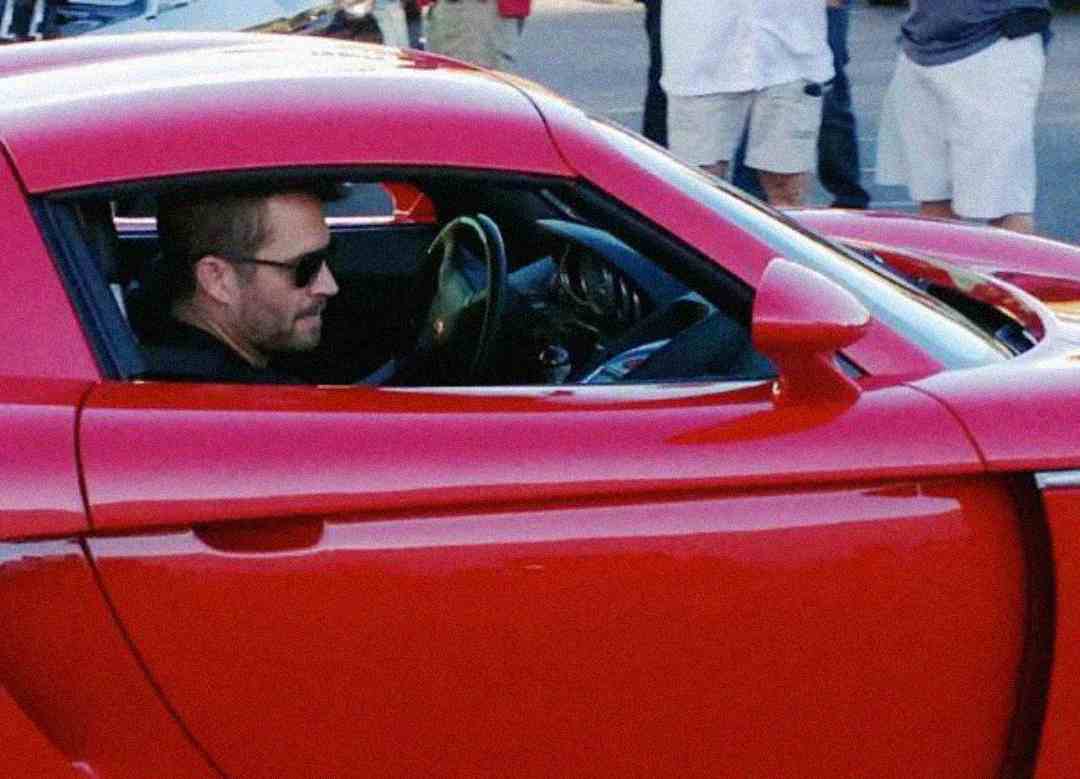 A man wearing sunglasses sits in the driver's seat of a bright red sports car with the window open. Several people are standing in the background.