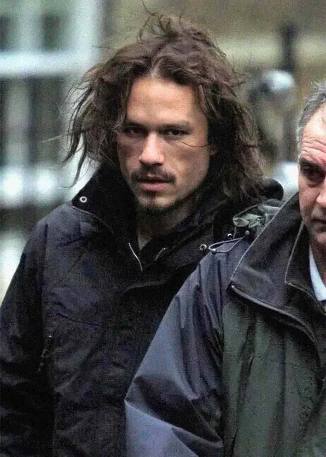 A man with long, tousled brown hair and facial hair wearing a dark jacket looks intensely at the camera while standing outdoors next to another man in a coat.
