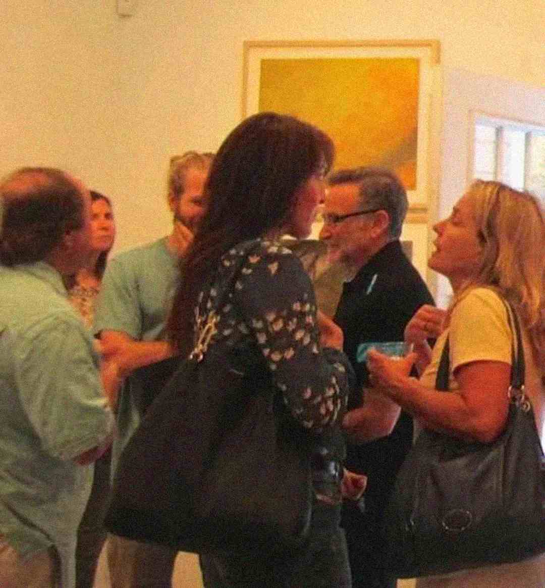 A group of people are standing and conversing in an art gallery with abstract artwork on the wall behind them. Some are holding drinks and wearing casual clothing and bags. The atmosphere appears lively and social.