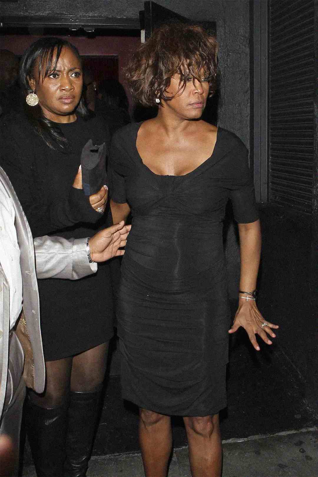 Two women in black dresses stand close together at night; one woman looks disheveled and is being supported by the other as they exit a building.