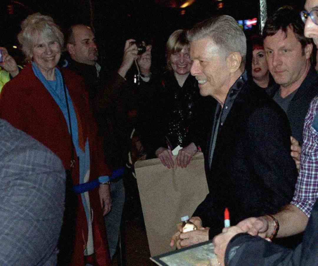 A man in a dark suit smiles while holding a pen and signing autographs for fans in a crowd at night. People around him are taking photos and watching him, and a woman in a red coat stands nearby, smiling.