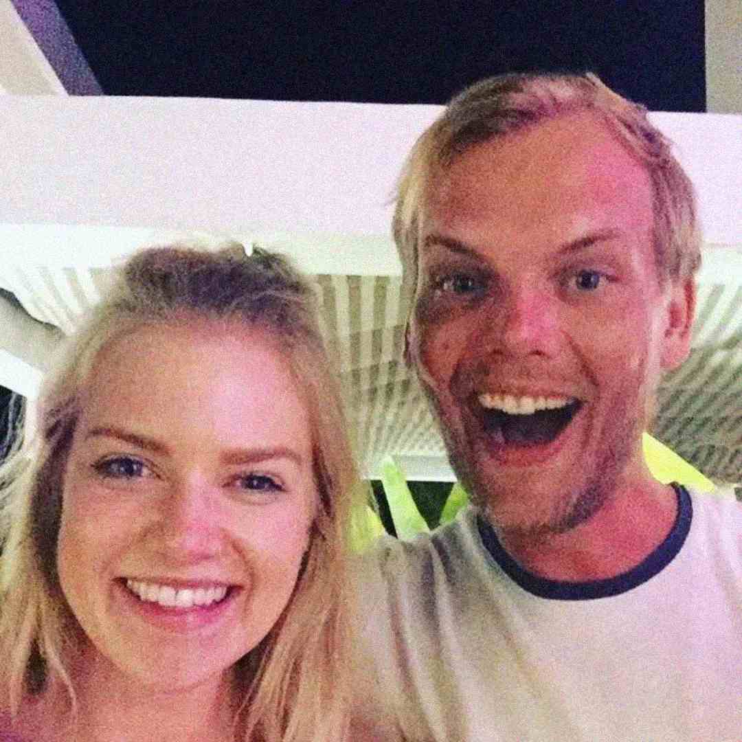 Two people with blonde hair are smiling at the camera. The person on the right has an open-mouthed, excited expression, while the person on the left has a relaxed smile. They appear to be outdoors at night.