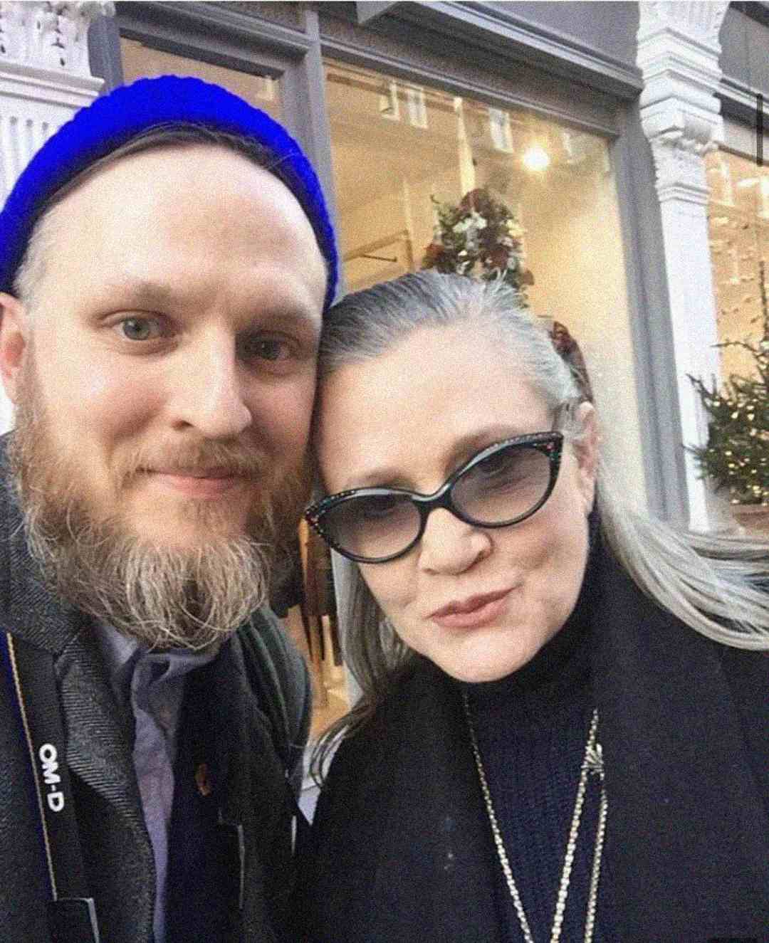 A bearded man in a blue beanie poses for a selfie with a woman wearing black cat-eye sunglasses and a dark coat in front of a building with large windows.