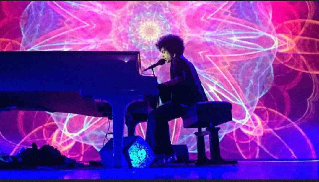 A person with curly hair plays a grand piano and sings into a microphone on stage, with a colorful, kaleidoscopic light display in the background.