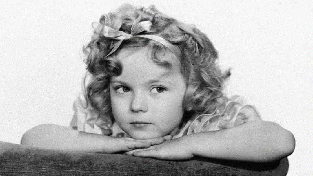 A young girl with curly blonde hair and a ribbon headband rests her chin on her folded arms, looking to the side with a thoughtful expression in a black and white photo.