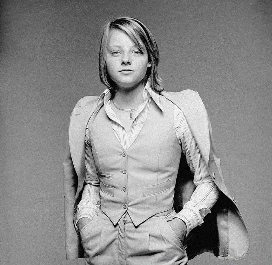 A young person with shoulder-length hair wears a light-colored suit vest over a striped shirt, with the suit jacket draped over their shoulders and hands in their pockets, posing confidently against a plain background.