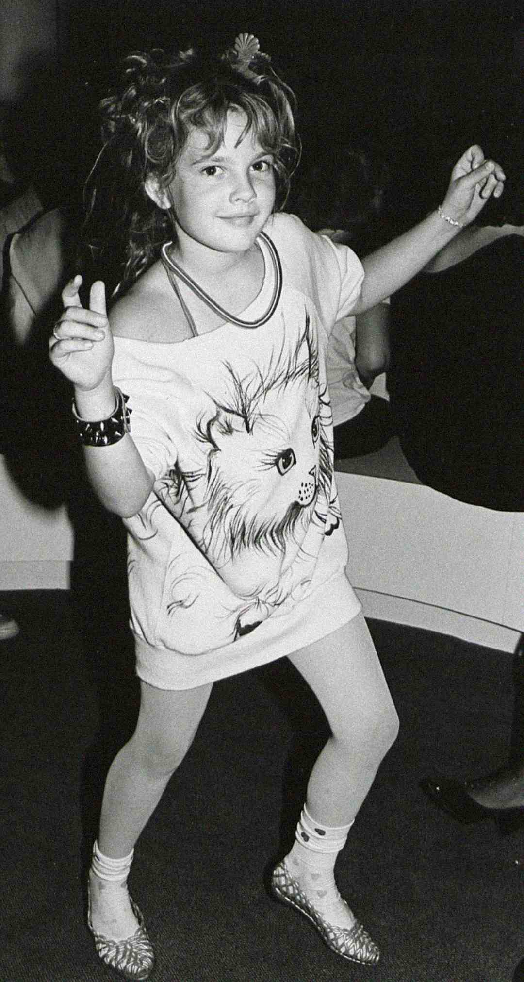 A young girl dances wearing a T-shirt with a large cat illustration, a bracelet, patterned socks, and flats. She smiles, with her hair styled in a messy, playful way. The photo is in black and white.