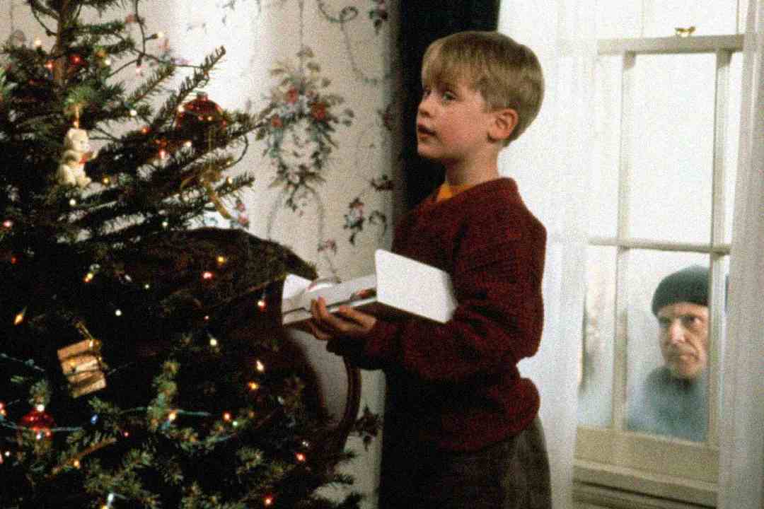A young boy in a red sweater stands by a decorated Christmas tree, holding a toy. Outside the window, a man in a dark hat and coat peers in at him.