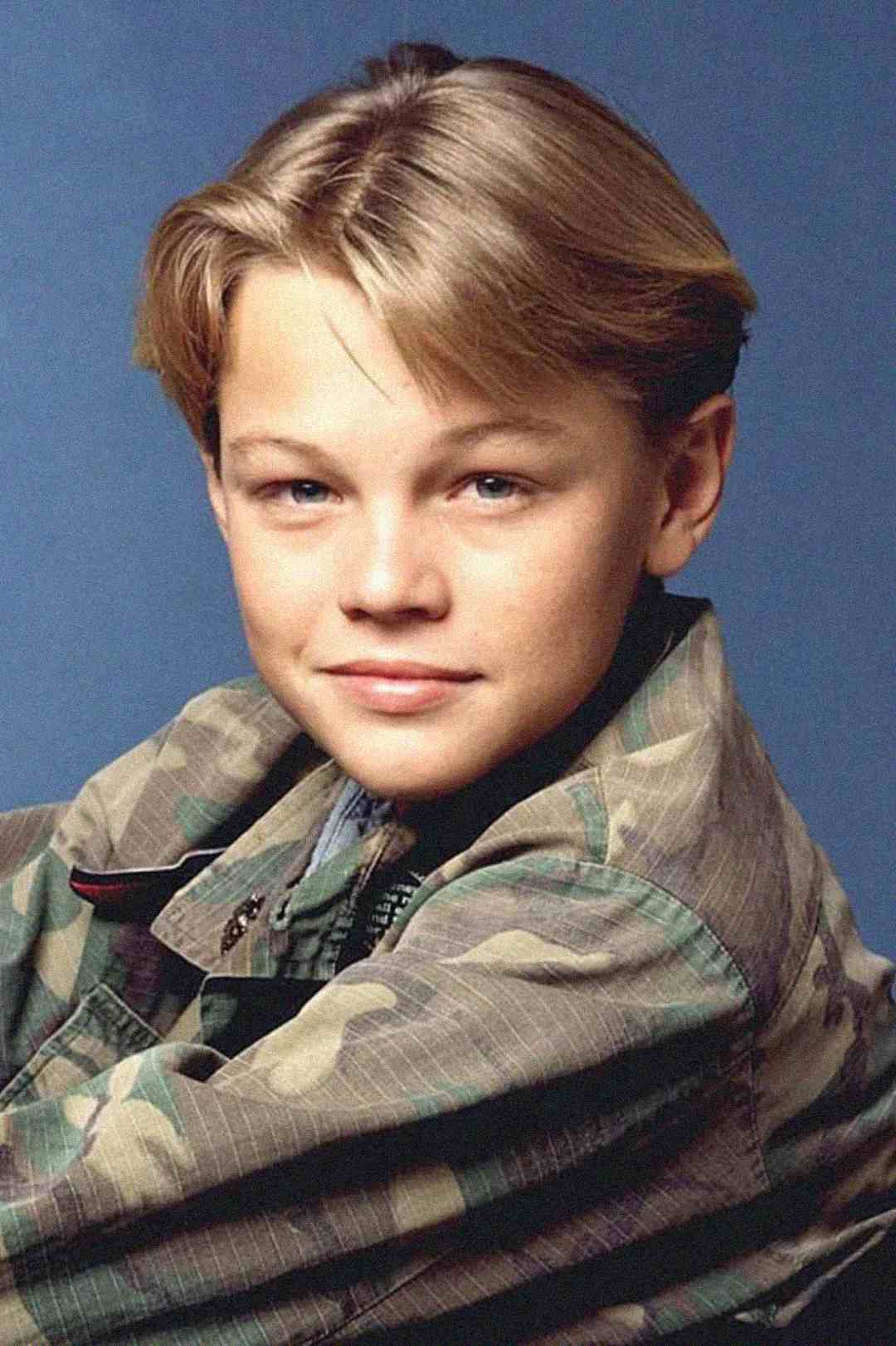 A young boy with light brown hair and blue eyes wears a camouflage jacket and smiles slightly, posing in front of a plain blue background.