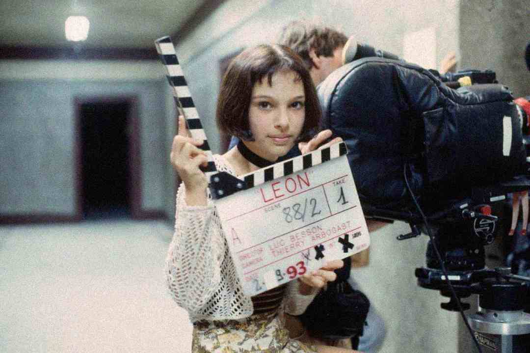 A young girl with short brown hair holds a film clapperboard labeled "LEON" near a film camera in a hallway, preparing for a movie scene.