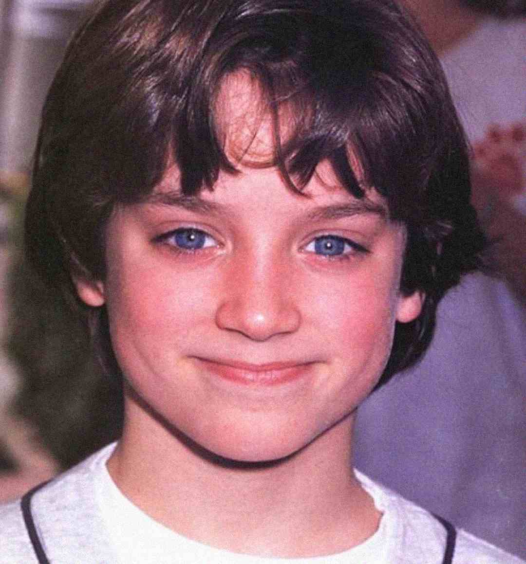 A young boy with medium-length brown hair and bright blue eyes smiles softly at the camera, wearing a white shirt with a dark collar. The background is softly blurred.
