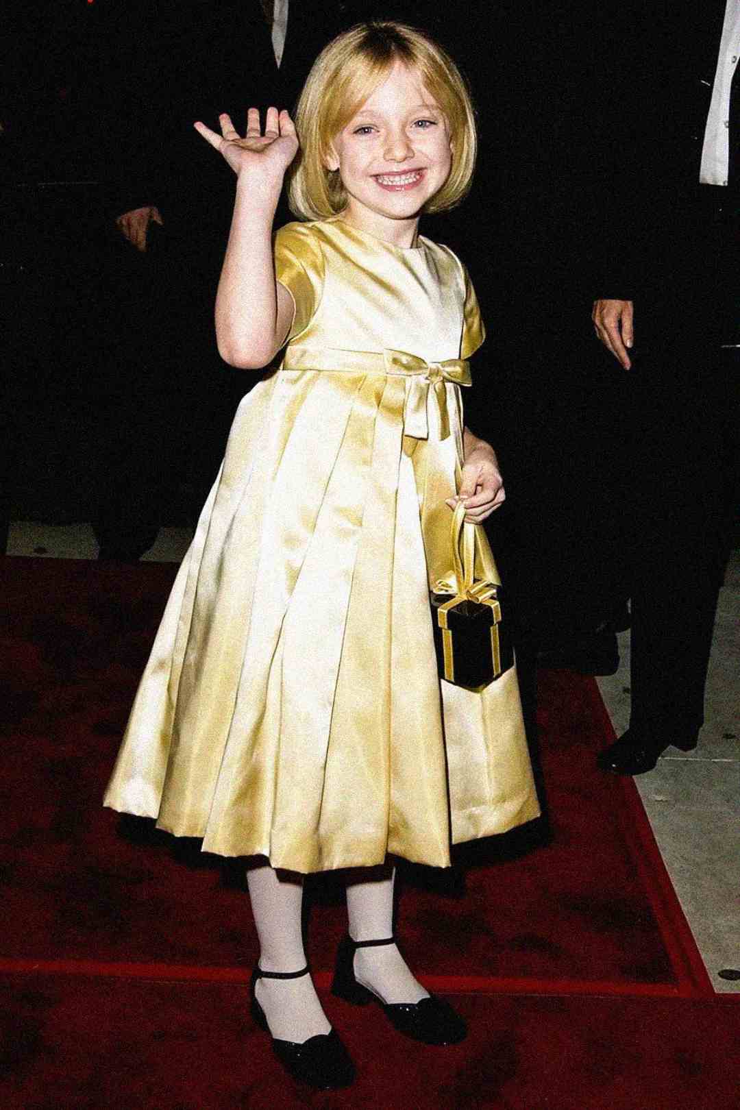 A young girl in a shiny gold dress with a matching purse smiles and waves, standing on a red carpet. She has short blonde hair, white tights, and black shoes. The background shows people in formal attire.