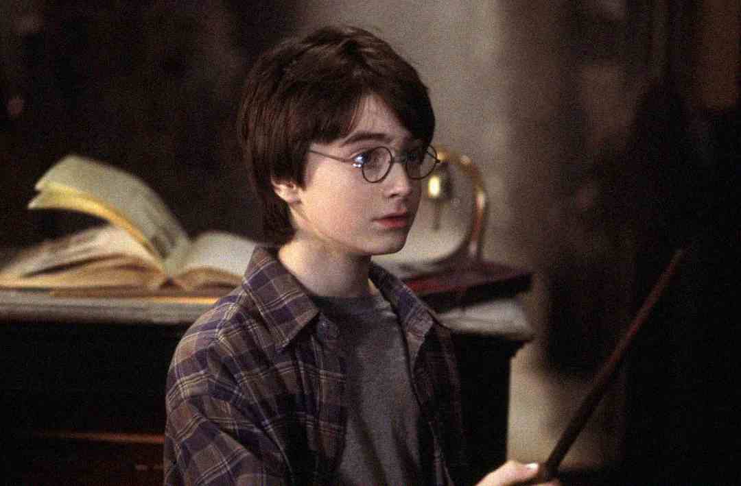 A young boy with round glasses and dark hair holds a wand, standing indoors with an open book and a glass orb in the background. He wears a plaid shirt over a t-shirt and looks focused.