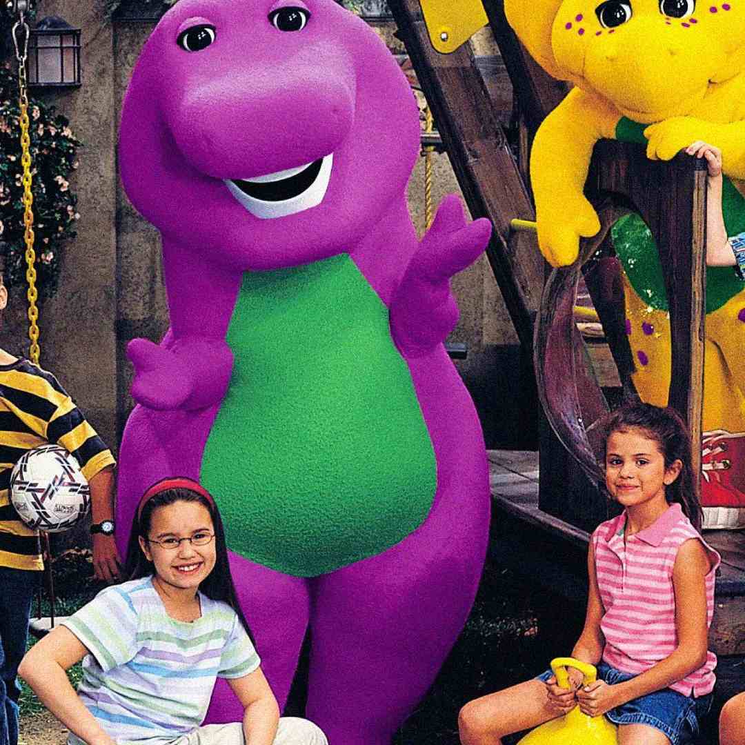 Barney the purple dinosaur stands and smiles with a yellow dinosaur and two young girls, one wearing glasses and a striped shirt, and the other in a pink top and shorts, in an outdoor play area.