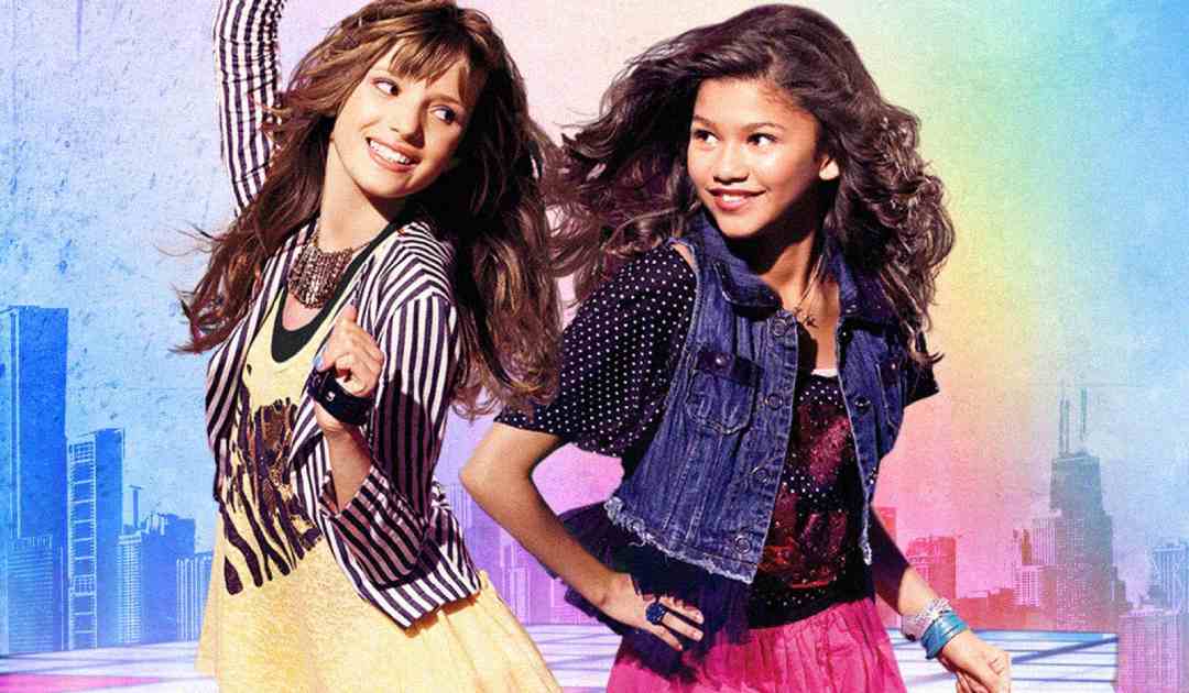 Two smiling teenage girls dance together against a colorful cityscape background. One wears a striped jacket and yellow top, the other wears a denim vest and pink skirt. Both have long hair and energetic poses.