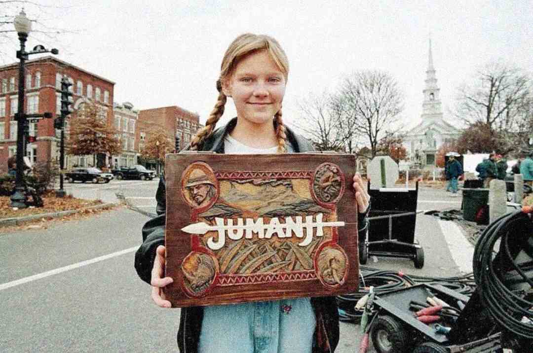 A young girl with blonde hair and a denim jacket smiles and holds up a wooden Jumanji board game prop on a street set, with film equipment and buildings visible in the background.