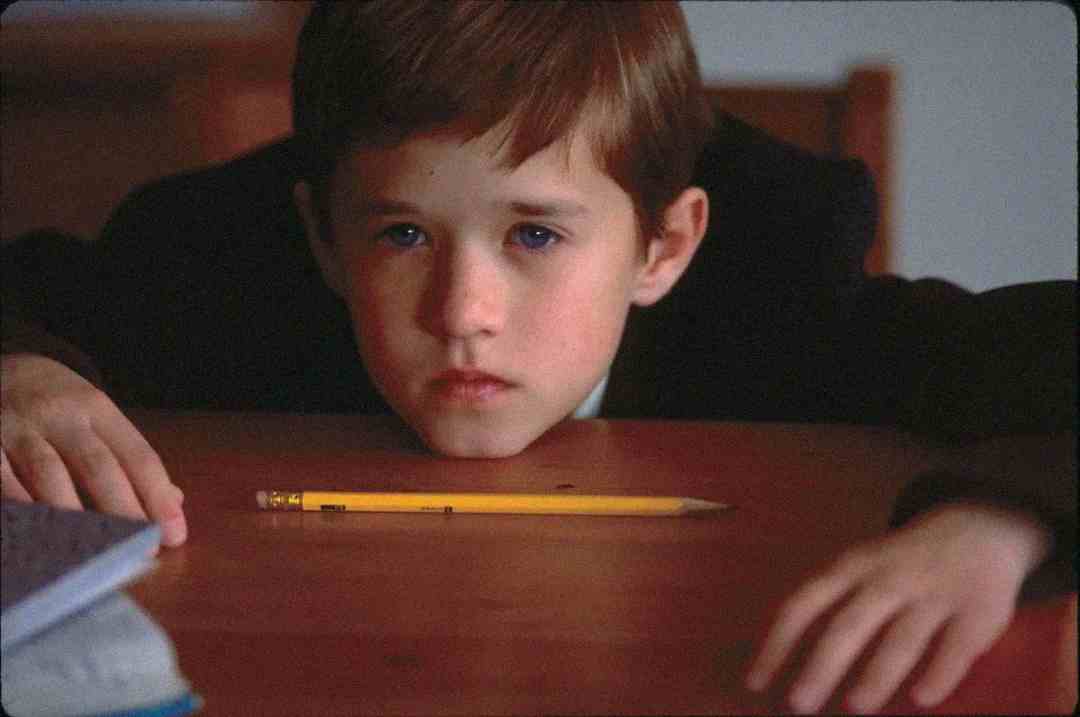 A young boy with short brown hair sits at a table, staring intently at a yellow pencil lying in front of him. His hands rest on either side of the table, and he appears deep in thought or concentration.