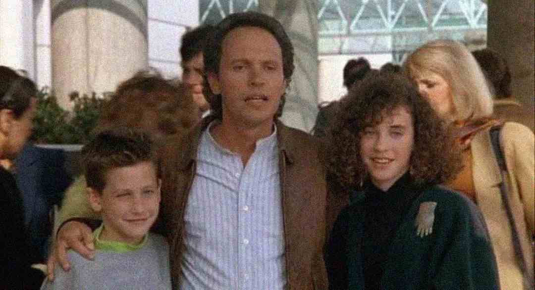 A man stands with his arms around a smiling boy and girl in an outdoor, crowded area. The group appears happy, with people and large columns visible in the background.