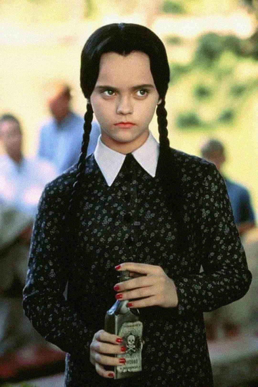 A girl with long dark braids, pale skin, and a serious expression wears a dark floral dress with a white collar. She holds a bottle with a skull label, standing outdoors with a blurred background.