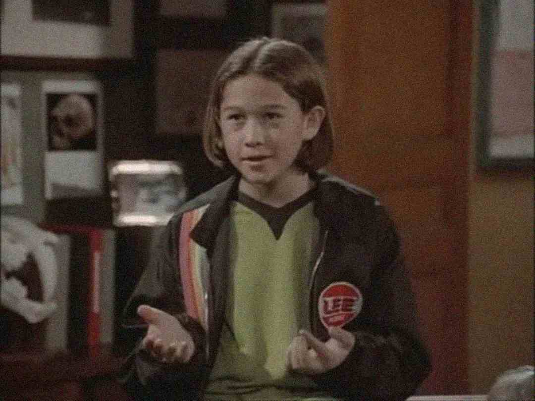 A young person with straight, shoulder-length brown hair wears a black jacket with colored stripes and a green shirt, standing indoors with hands open, appearing to speak or gesture.