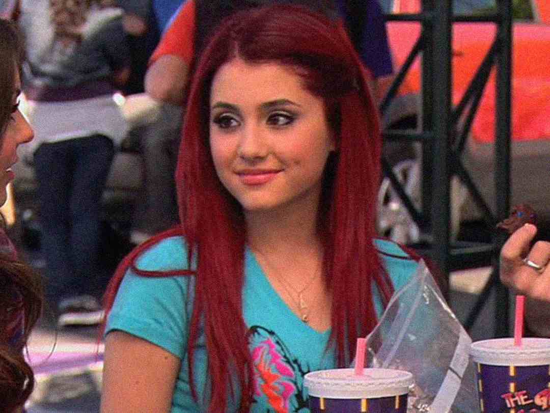 A young woman with long, bright red hair sits outdoors at a table, wearing a blue T-shirt. She is smiling slightly, with drinks and snacks in front of her, and people are visible in the blurred background.