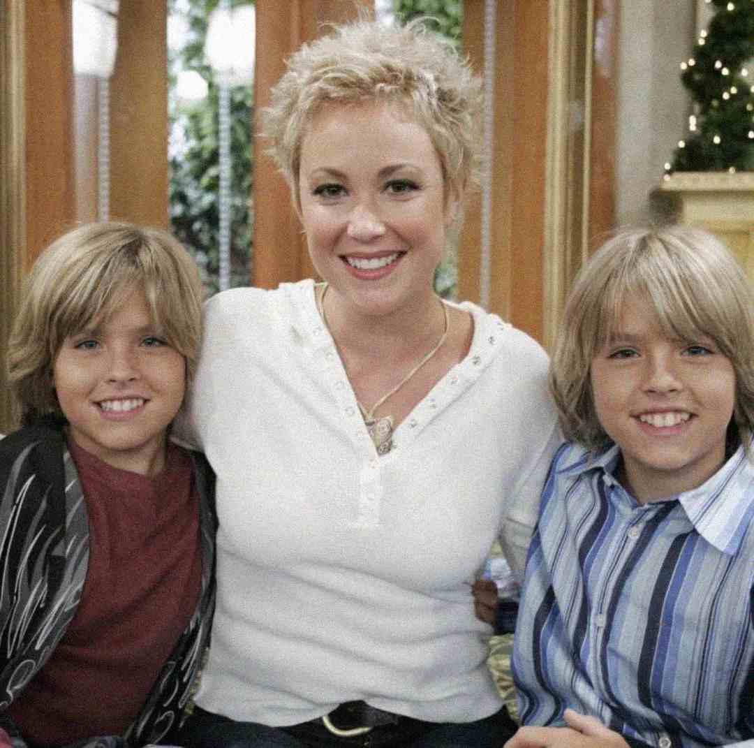 A woman with short blonde hair sits between two smiling blonde boys, all looking at the camera indoors with a decorated background.