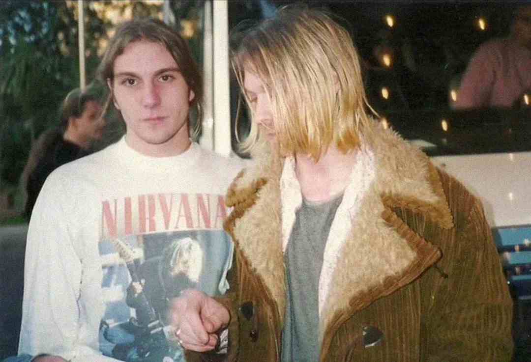 Two young men stand together. One wears a white Nirvana shirt, looking at the camera; the other has shoulder-length blond hair and wears a brown shearling coat, looking down and holding the first man's hand.