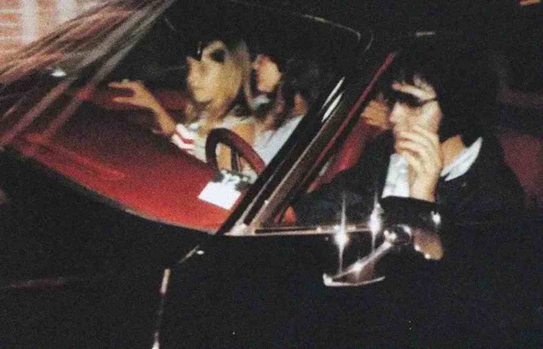 Three people sit in a car; the driver wears dark sunglasses and sideburns, while two passengers sit beside and behind him. The photo is slightly blurry and taken from outside the vehicle.