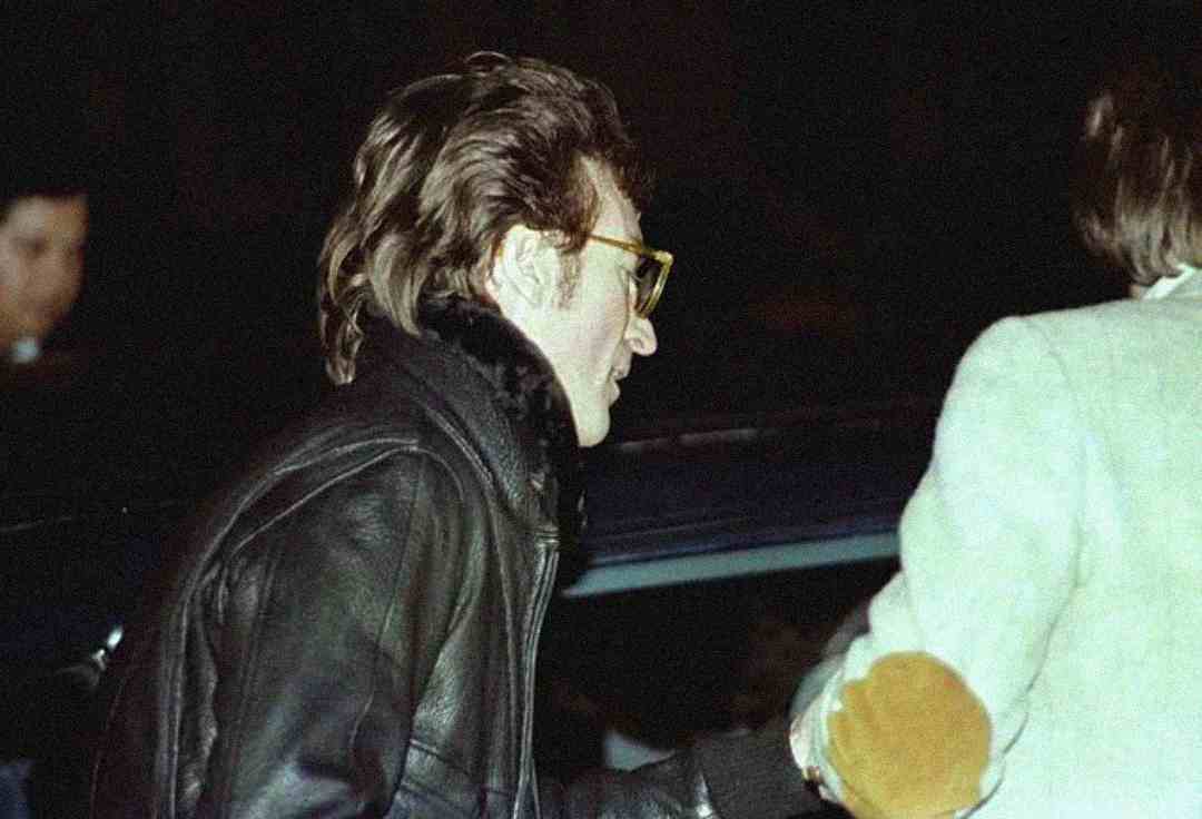 A person with shaggy hair, wearing yellow-framed glasses and a black leather jacket, stands next to another person in a light-colored coat near a car at night.