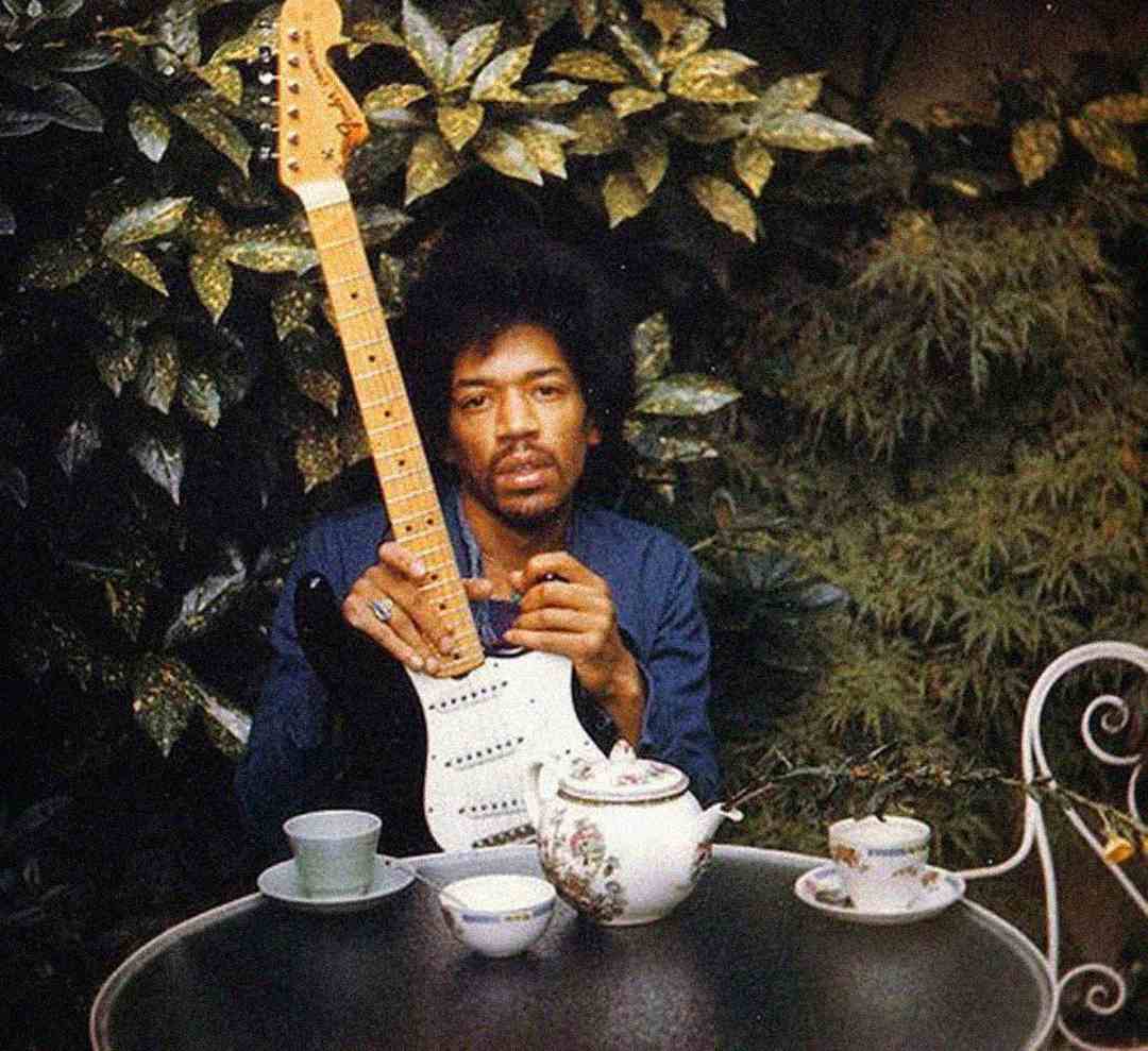 A man with an afro hairstyle sits at an outdoor table set for tea, holding a white electric guitar upright. The table has a teapot and cups, and lush green foliage fills the background.