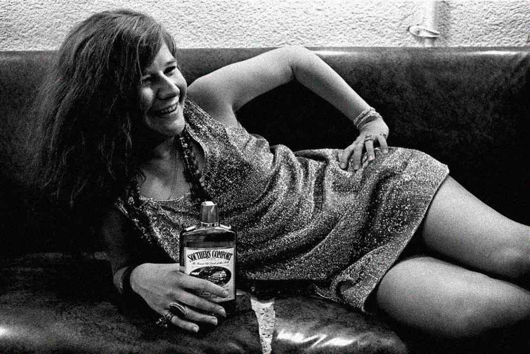 A woman in a glittery dress reclines on a leather couch, smiling and holding a large bottle labeled “Southern Comfort.” The photo is black and white, and the scene appears relaxed and candid.