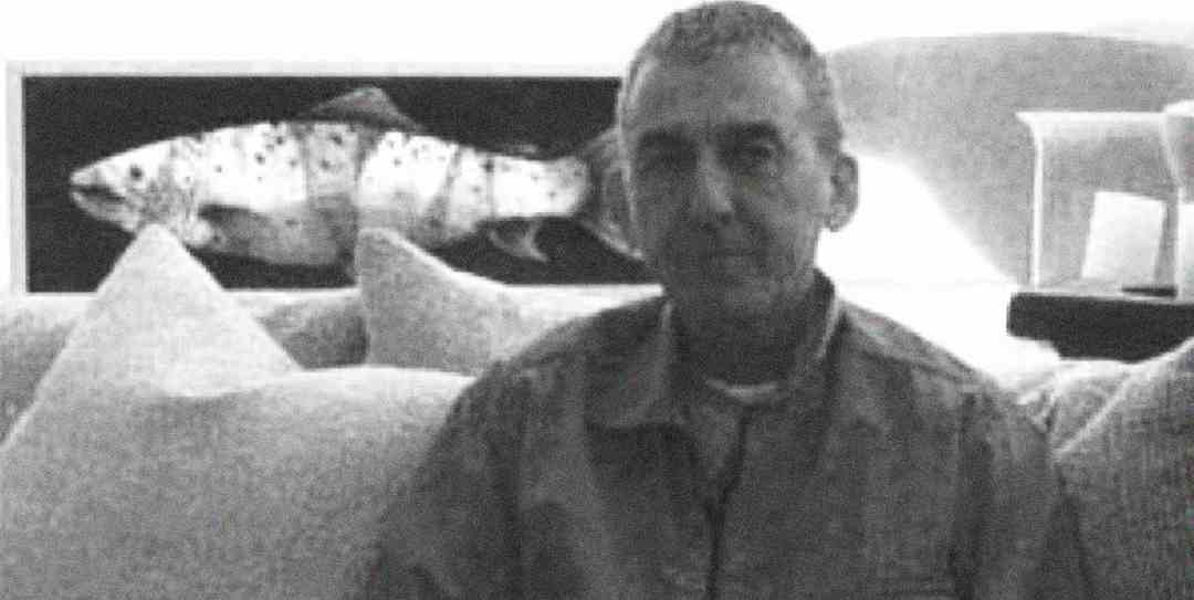 A man sits on a couch in front of a framed picture of a fish. The image is in black and white, and the man is looking toward the camera with a neutral expression. Several pillows are visible around him.
