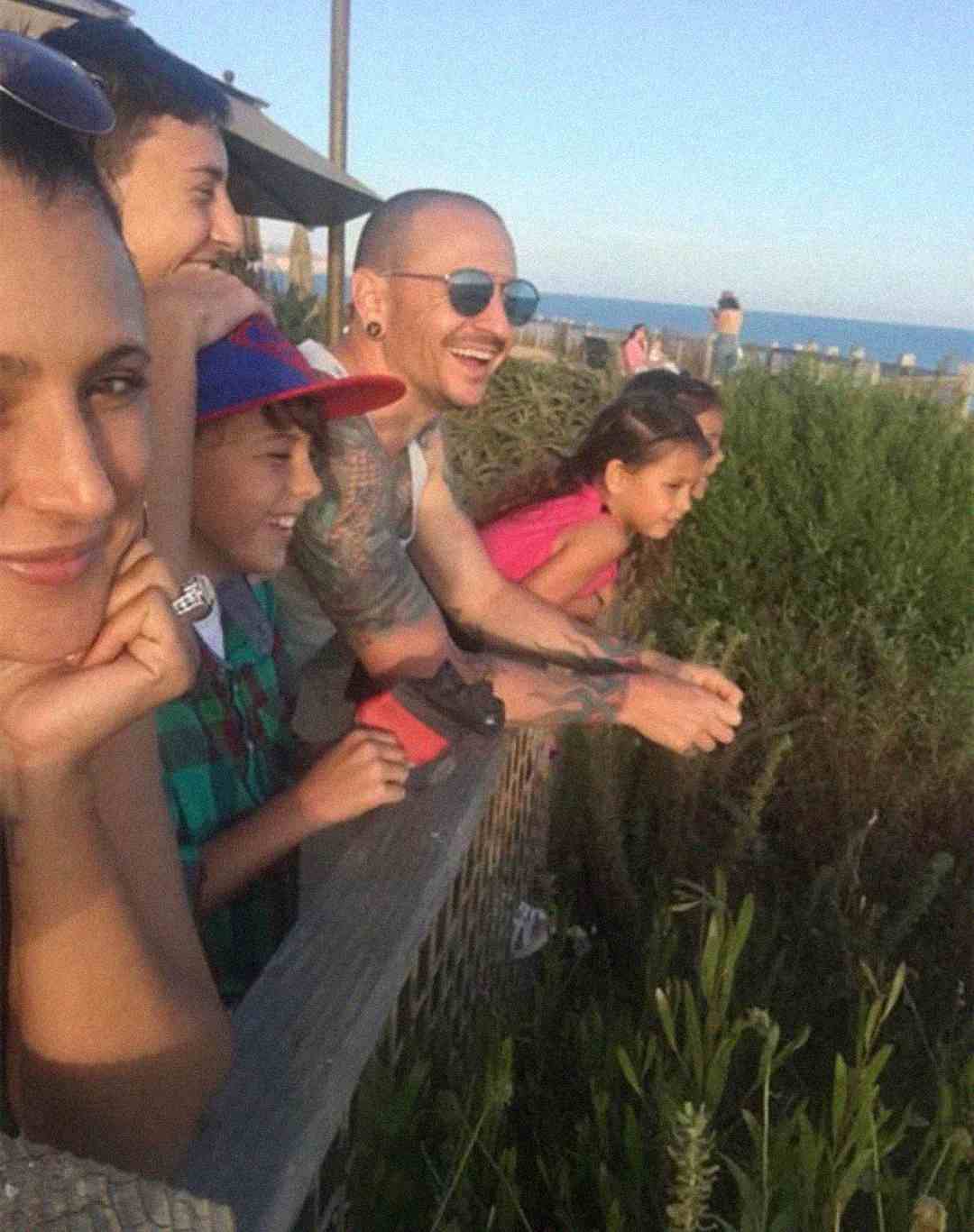A group of smiling people, including adults and children, stand together outdoors by a wooden fence, enjoying a sunny day with greenery and the ocean in the background.