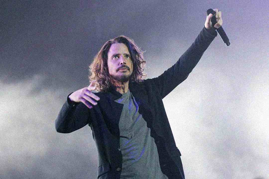 A man with long hair holds a microphone in one hand and raises his other arm while performing on stage, with a smoky, purple-toned background.