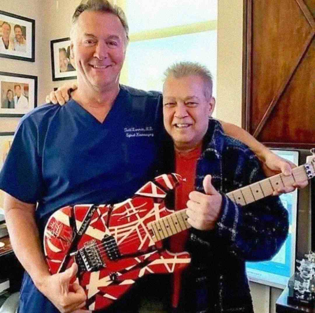 Two men smiling for a photo indoors; one is wearing blue scrubs, and the other holds a red, white, and black striped electric guitar while wearing a plaid shirt. Framed photos and a computer are visible in the background.