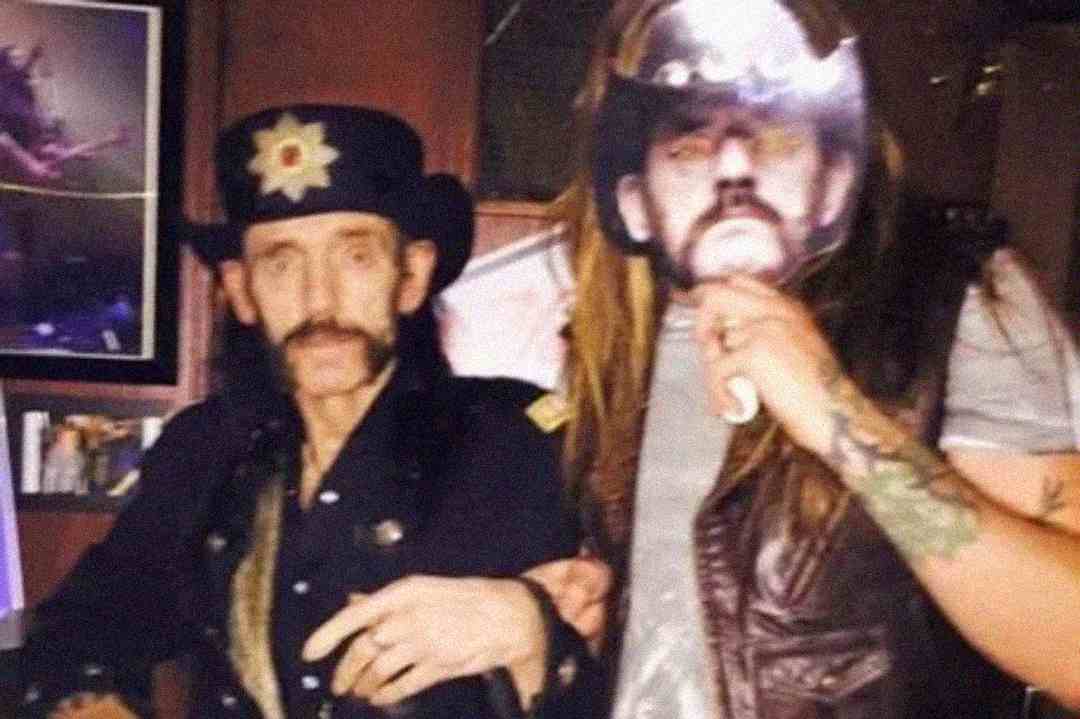Two people pose together; one wears a black hat and mustache, while the other holds up a paper mask of the first person's face, creating a humorous effect. The setting appears to be indoors with framed pictures in the background.