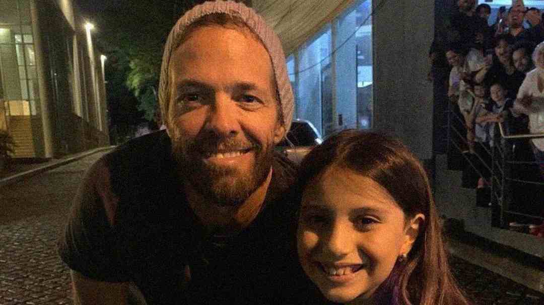 A man wearing a knit beanie and a dark shirt smiles beside a young girl with long brown hair. They are outdoors at night with a crowd of people and a modern building in the background.