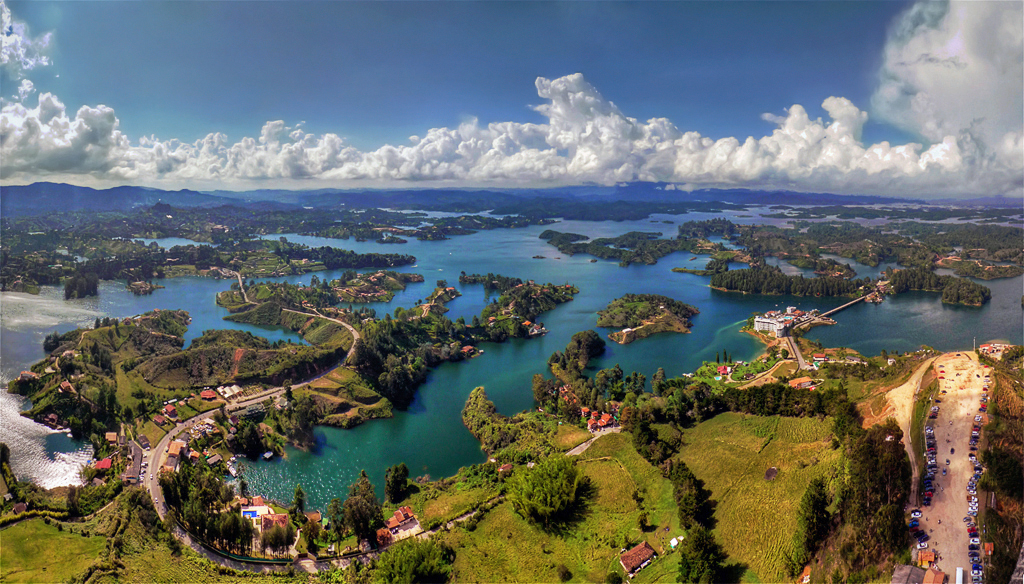 A panoramic aerial view of a lush, green landscape dotted with houses and winding roads, surrounded by numerous blue lakes and islands under a partly cloudy sky.