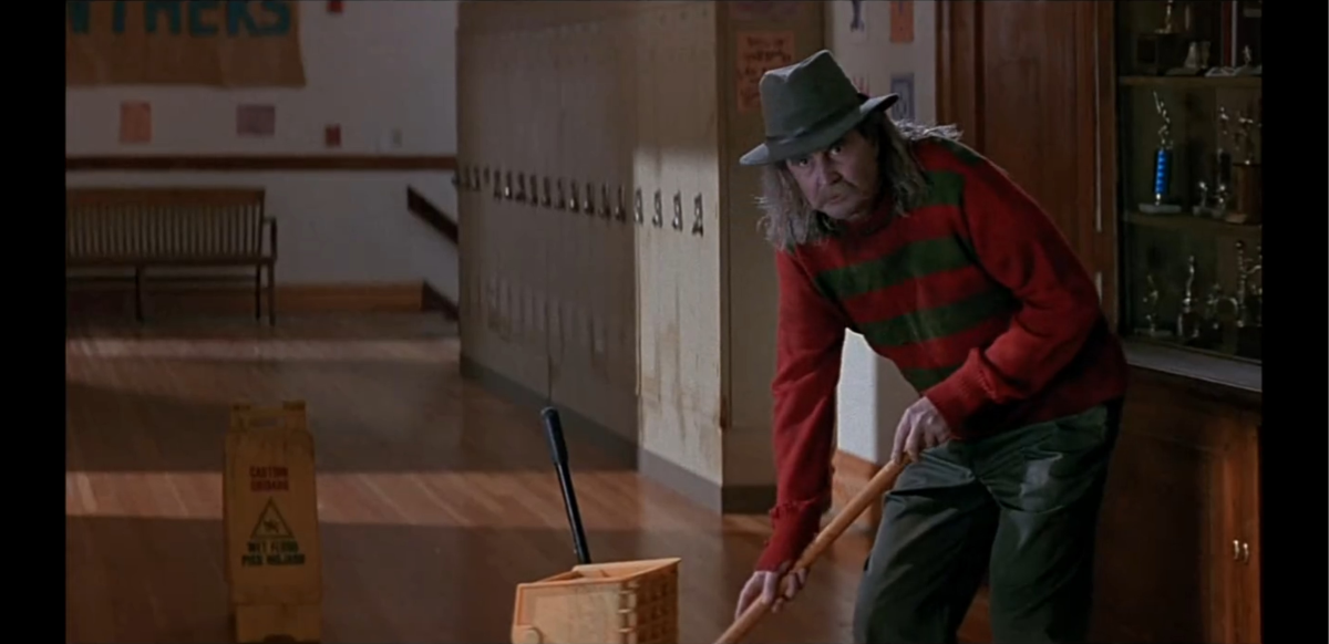 A man with long gray hair, wearing a fedora and a red-and-green striped sweater, mops the floor in a school hallway lined with lockers. A yellow caution sign and mop bucket are nearby.