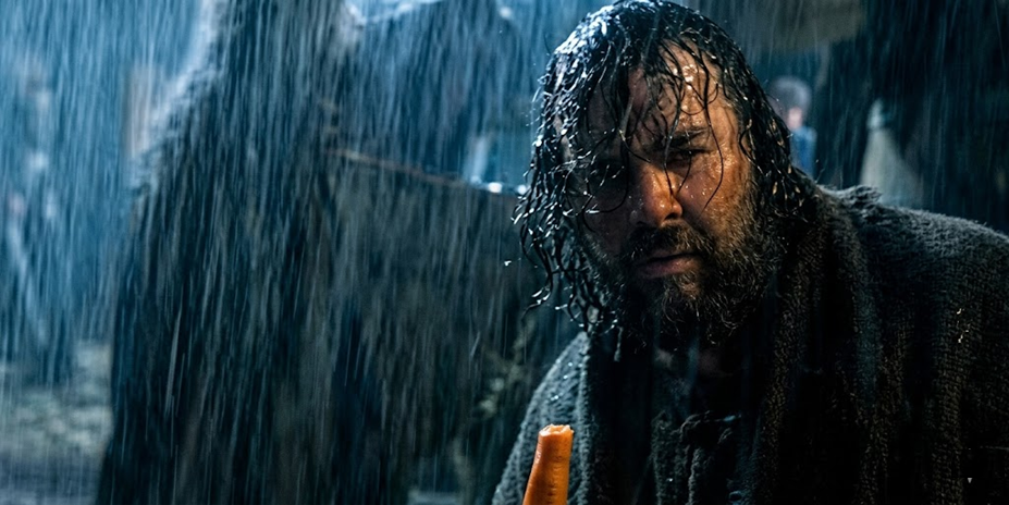 A man with a beard and wet hair, wearing dark clothes, holds a carrot in the pouring rain.