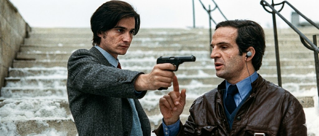 A man in a grey suit points a gun at another man wearing a brown leather jacket and an earpiece, on snowy stairs.
