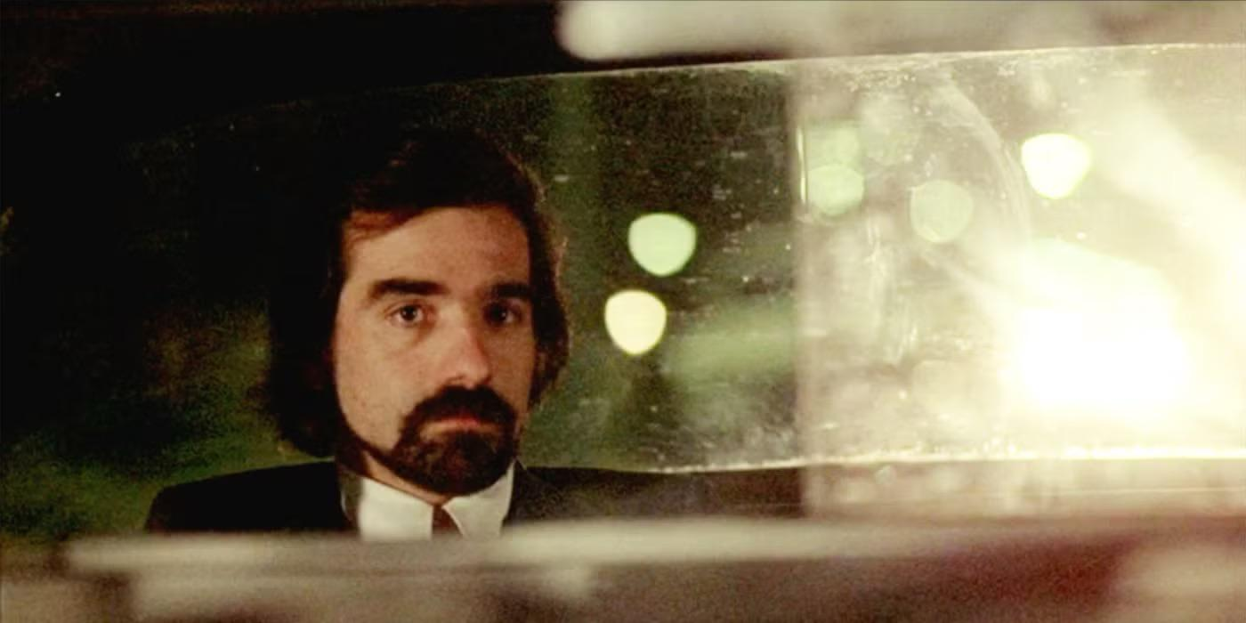 A man with dark hair and a beard is seen in the rearview mirror of a car at night, wearing a white shirt and dark jacket, with blurred lights in the background.